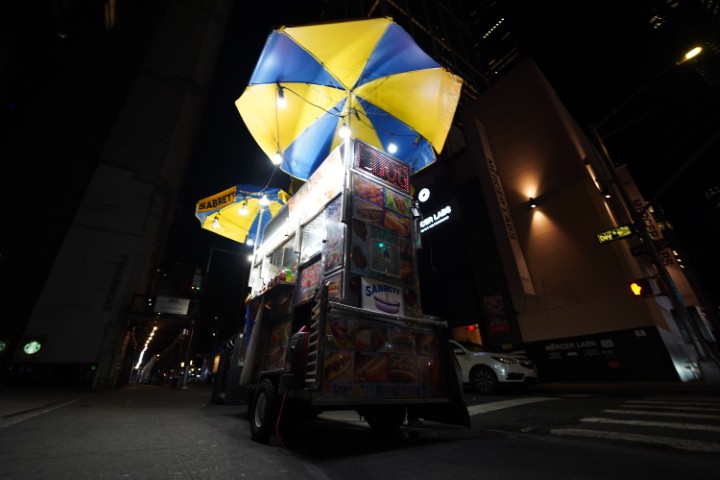 A hot-dog stand in a street of NYC, at night, taken from a low angle, with two bright blue and yellow umbrellas above.